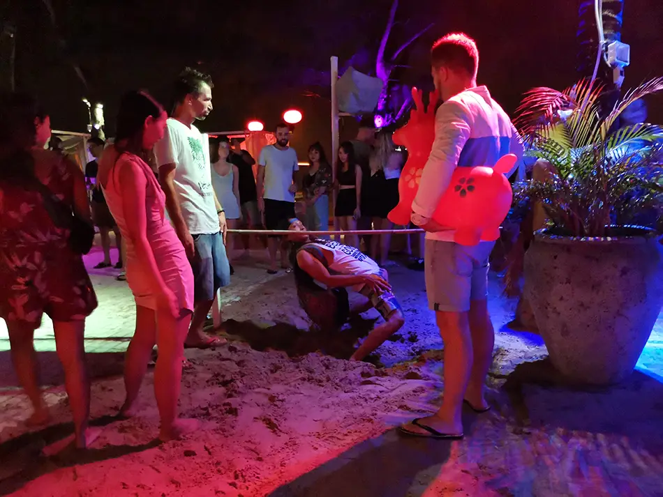 Backpackers dance on sand under red-blue lights during hostel party in Vietnam.