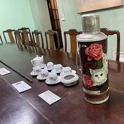 Long wooden table with chairs, porcelain tea cups, and preserved objects displayed in a bunker room.