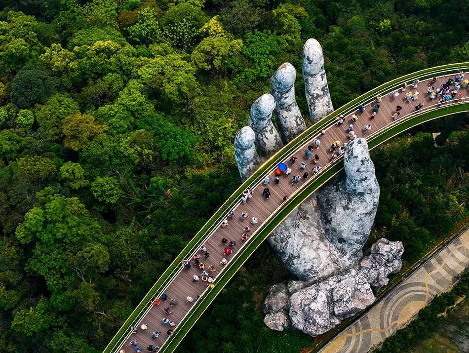 Pedestrian bridge supported by giant stone hands stretches above forested mountains.