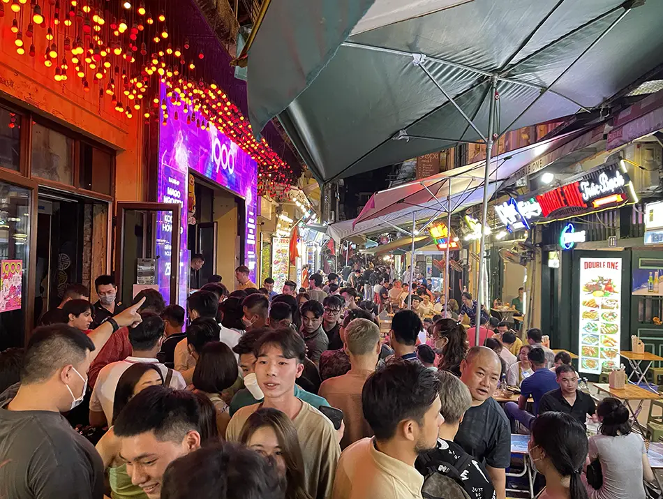 Packed alley filled with street tables, neon signs, and crowds enjoying nightlife in hanoi after dark.