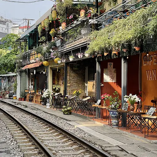 Railway tracks pass inches from small cafés with plants, tables, and hanging lanterns along the line.
