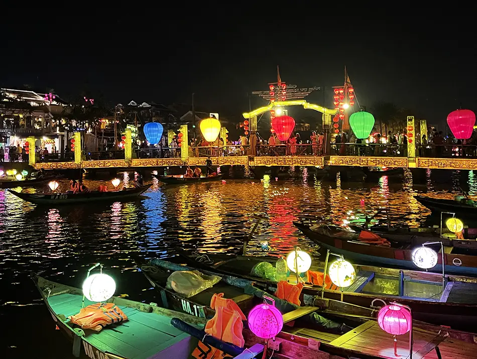 Lantern-lit boats float along a quiet river beside historic buildings after sunset.
