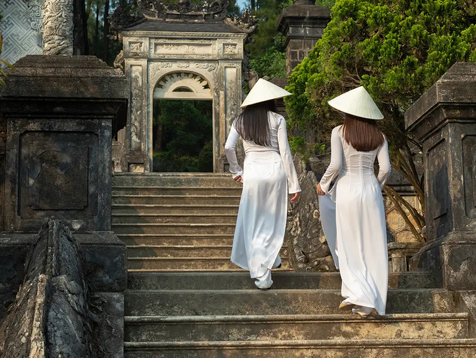 Two women in traditional dress walk stone steps near royal tombs