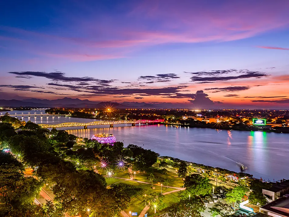 Illuminated riverbanks and bridges reflect on water showing nightlife in Hue.