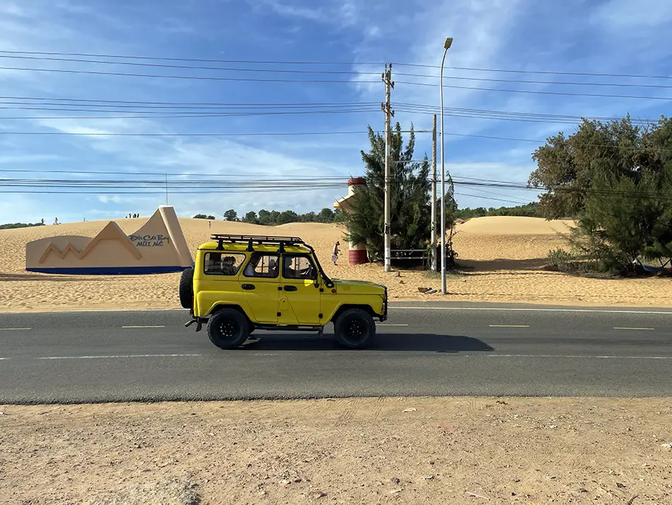 Yellow jeep driving past red sand dunes, showing activities in Mui Ne along the coastal road.