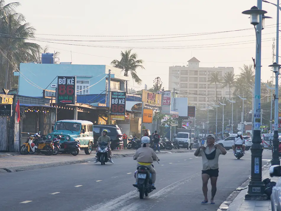 Daytime street scene with motorbikes, pedestrians, and local shops along the main coastal road.