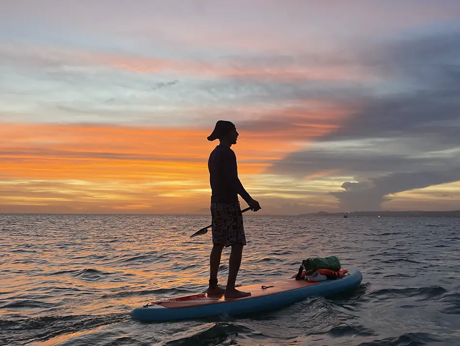 Traveler standing on a paddleboard at sea during sunset with colorful clouds on the horizon.