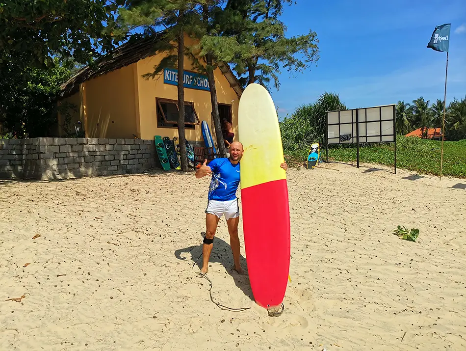 Traveler stands with red surfboard near a beach hut, windblown sand and blue sky.