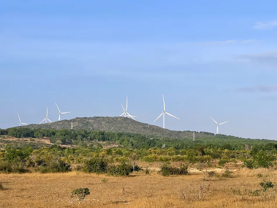 Wind turbines lined along a low hillside above dry fields and scattered greenery.