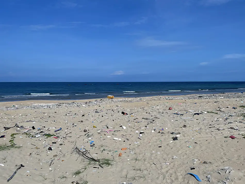 Plastic bottles and wrappers scattered across a sandy shoreline beside calm blue water.