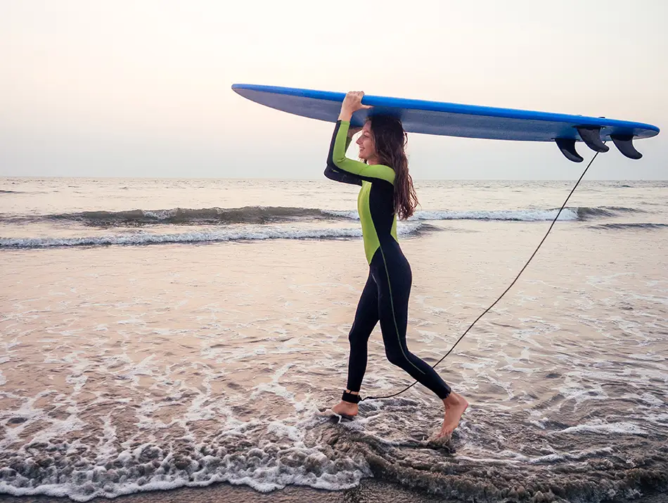 Surfer carries a longboard across a glassy tidal flat at sunrise, reflections shimmering.