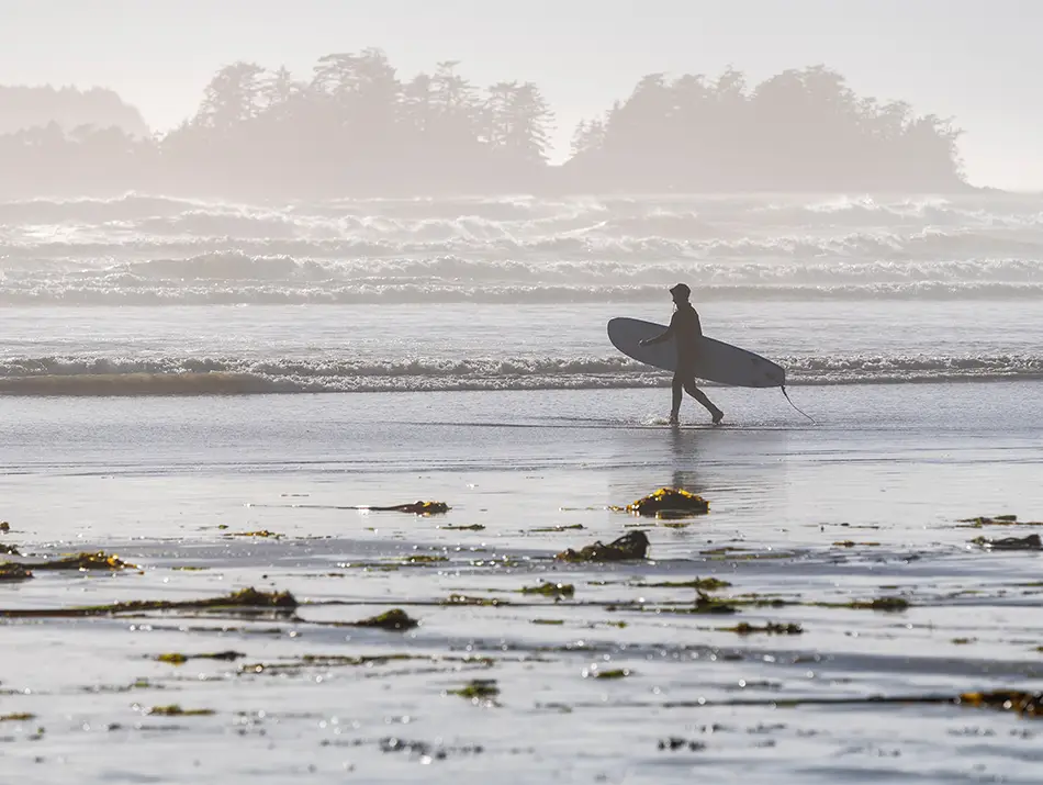 Surfer walks across misty Long Beach in Tofino, Canada, heading to the waves.