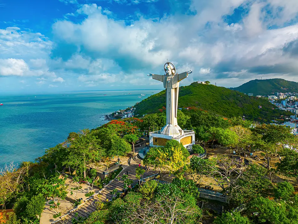 Large Christ the King statue standing on a green hillside in Vung Tau, overlooking the coastline and blue sea under a partly cloudy sky.
