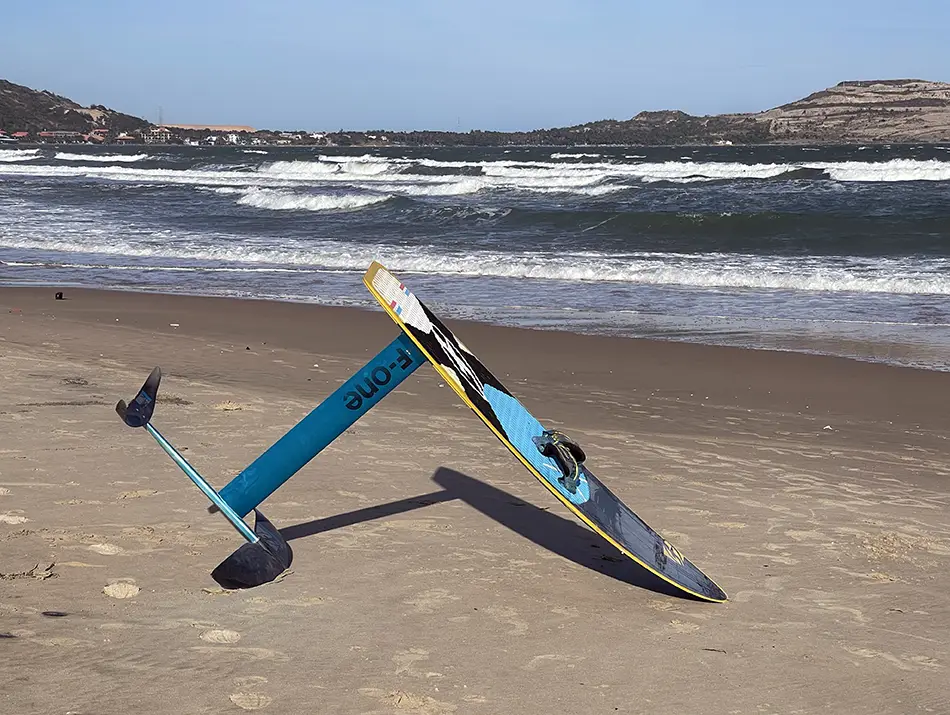 A hydrofoil kiteboard rests on wet sand beside breaking shore waves.