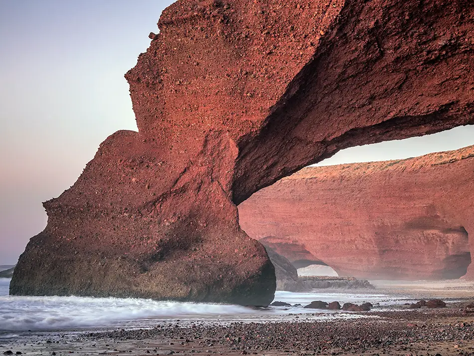 Impressive natural rock arches rise from the sandy beach against the ocean waves.
