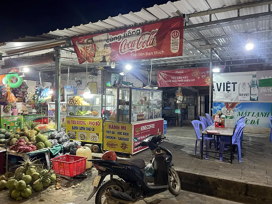 Nighttime street view with food stalls, signage, and motorbikes parked nearby.