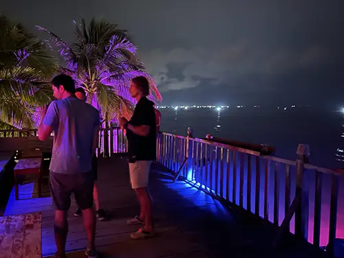 Tourists on a seafront balcony with colored lights overlooking the dark shoreline.