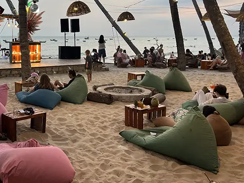 Bean bags and low tables arranged on the sand beneath sail shades.