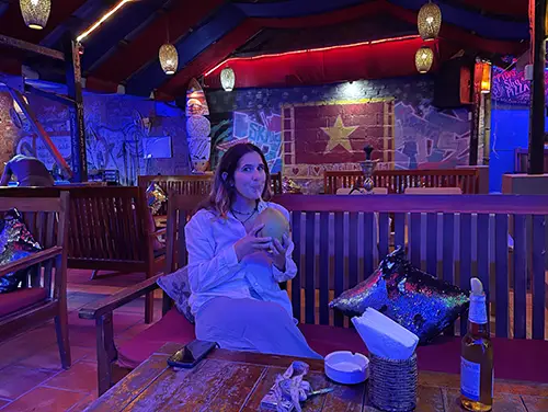 Traveler seated at a wooden terrace table under colored lights at night.