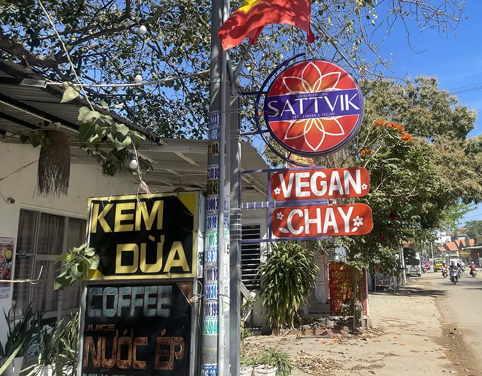 Roadside hanging sign marking vegan restaurantsin Mui Ne beside a small shop.