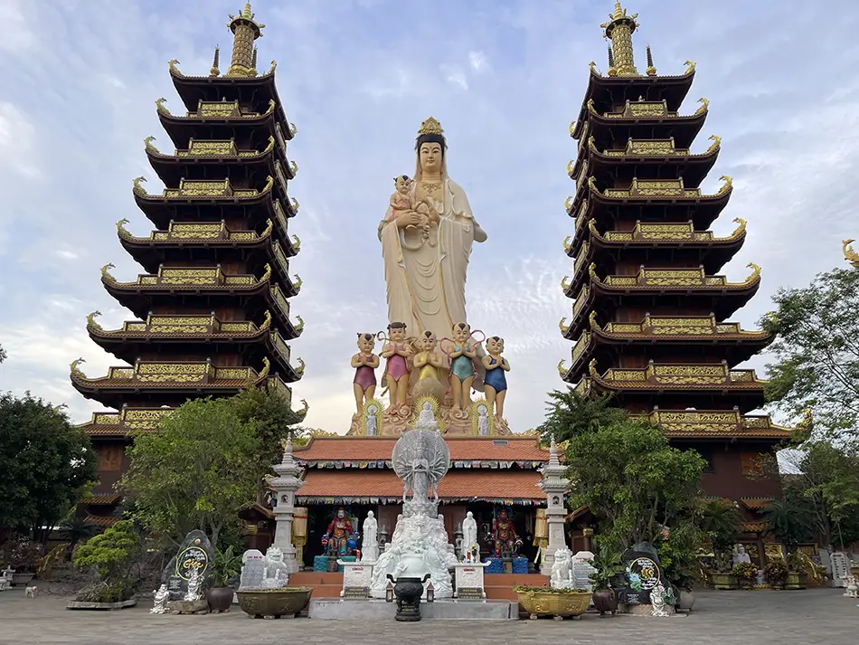 Twin multi-tiered Buddhist pagoda towers with a large white Guanyin statue between them at a temple in Mui Ne, Vietnam, set against a partly cloudy sky