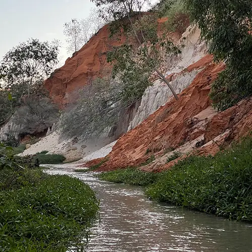 Red and orange eroded sandstone cliffs flanking the Fairy Stream in Mui Ne, with dense green vegetation growing along the top edge