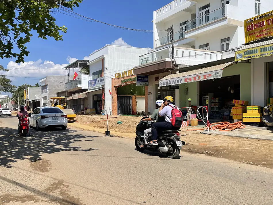 A quiet stretch of Mui Ne's main road lined with low-rise shophouses, parked motorbikes, and lush trees, giving a glimpse of everyday local life in the coastal town