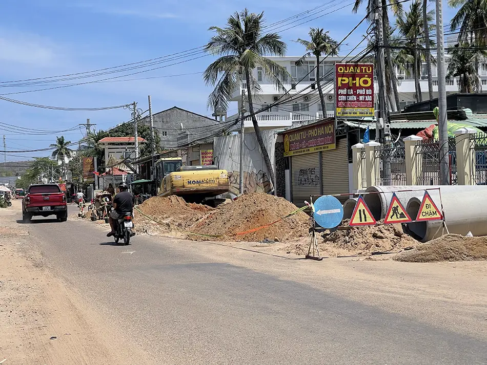 Road construction in Mui Ne with motorbikes passing warning signs along a dusty street.