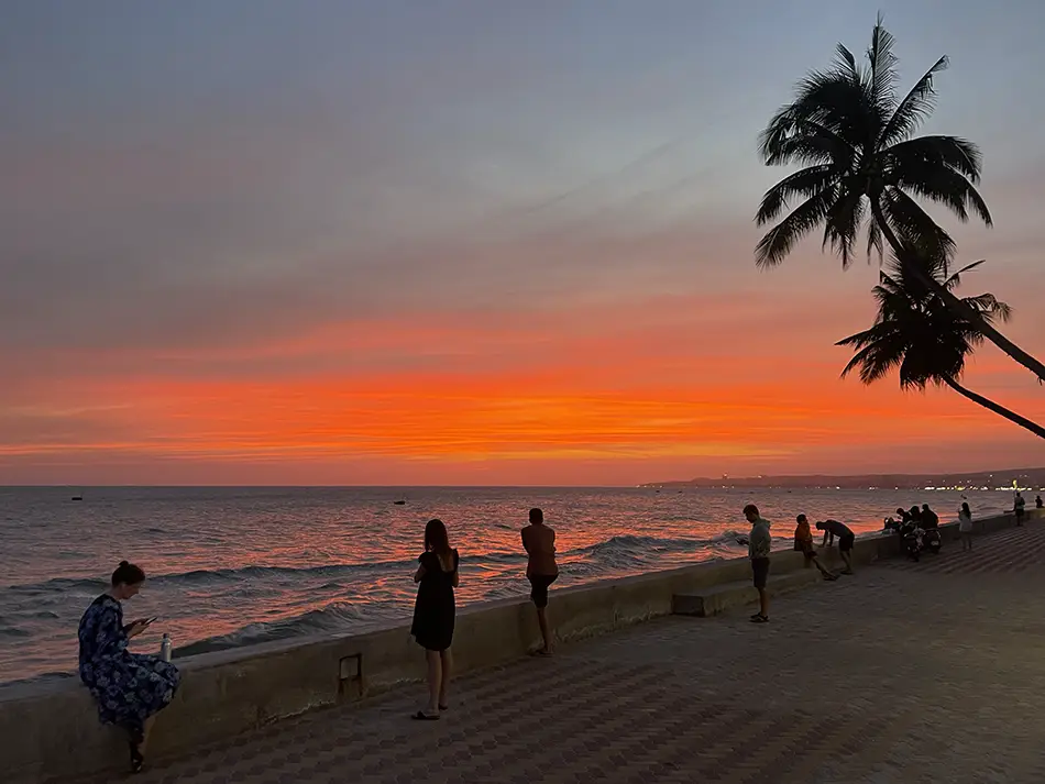 Sunset over Mui Ne beach with palm tree silhouette and travelers walking along the shoreline.