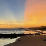Sunset over a sandy beach with palm trees, waves, and a coastal road in the distance