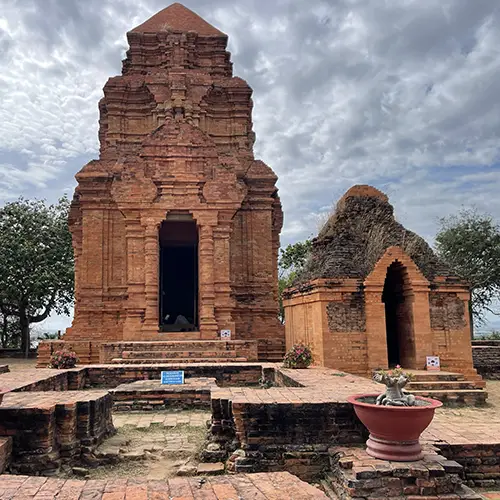 Ancient red-brick Cham tower temple rising against a cloudy blue sky in Phan Thiet near Mui Ne, with a ceremonial urn at its base — a key cultural landmark on any Mui Ne itinerary