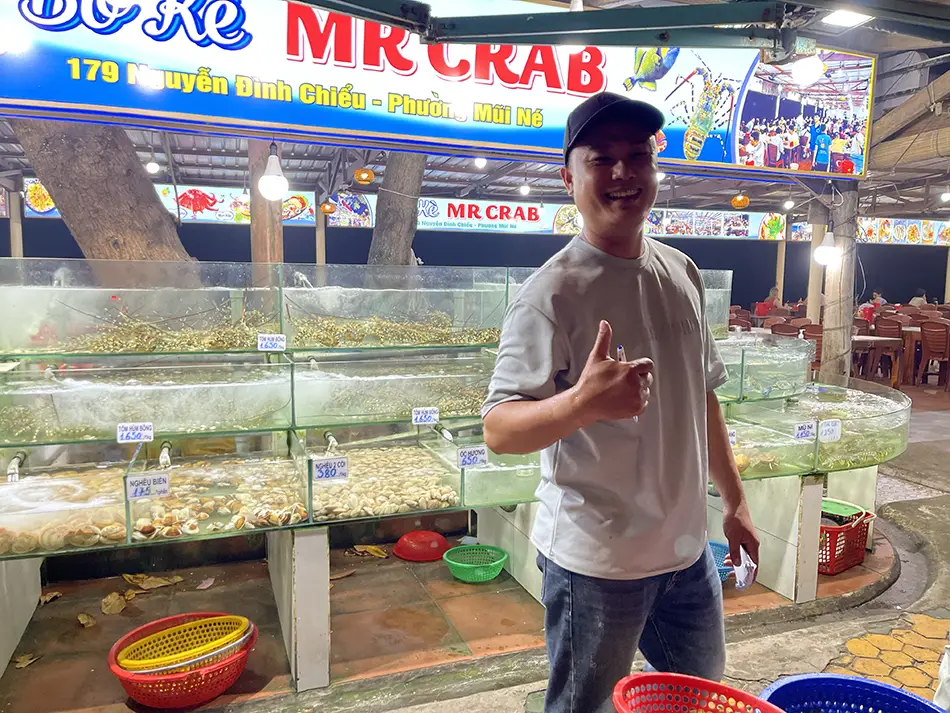 Seafood vendor standing in front of seafood tanks displaying Mui Ne dining options inside a bright hall.