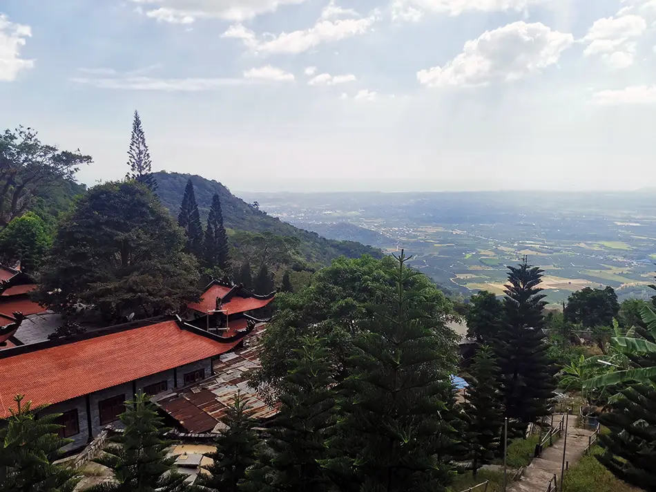 View from Ta Cu Mountain looking down over red-roofed pagoda buildings surrounded by dense jungle canopy, with a coastal valley and hazy horizon in the distance