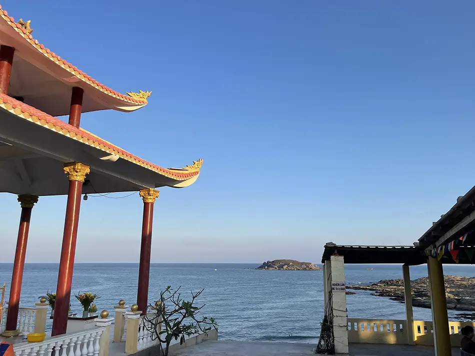 Traditional red-roofed Vietnamese shrine pavilion on a rocky coastal promontory overlooking a calm blue sea and distant islands on a clear day