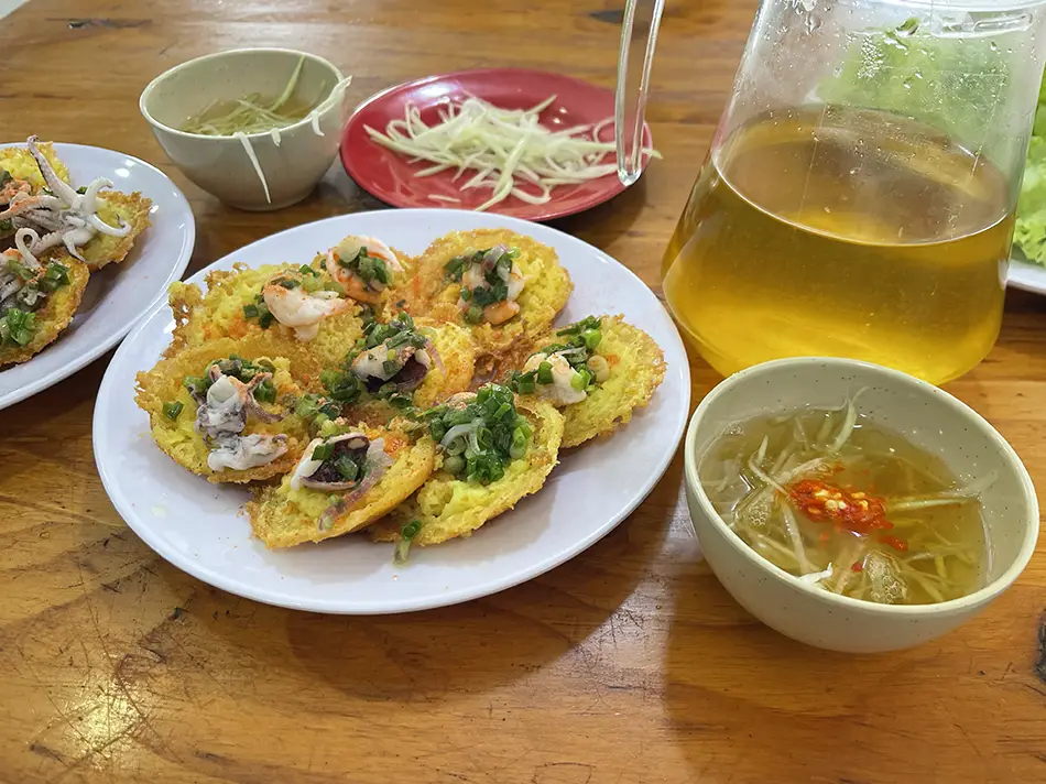 Plate of bánh khọt mini rice pancakes topped with shrimp and scallions, served with fresh herbs and dipping sauce
