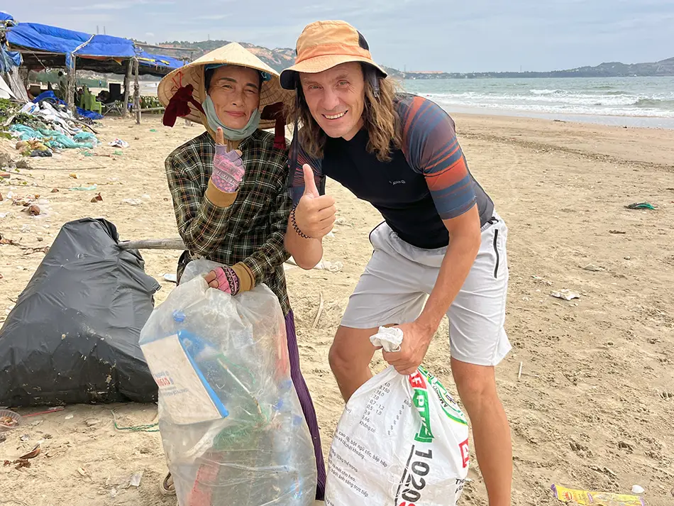 A man and a Vietnamese woman in a traditional conical hat giving a thumbs up while holding bags of collected plastic waste on a littered beach in Mui Ne