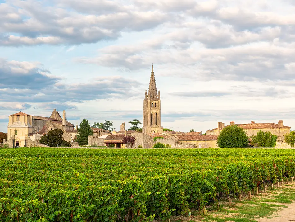 Green vineyard rows stretching toward the medieval church and stone village in the Bordeaux wine region, France, under a partly cloudy sky