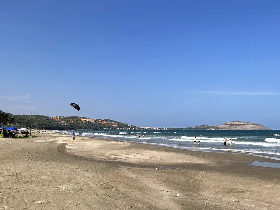 Wide sandy beach on the eastern side of the Mui Ne cape with kite lines visible in the sky and green hills in the background