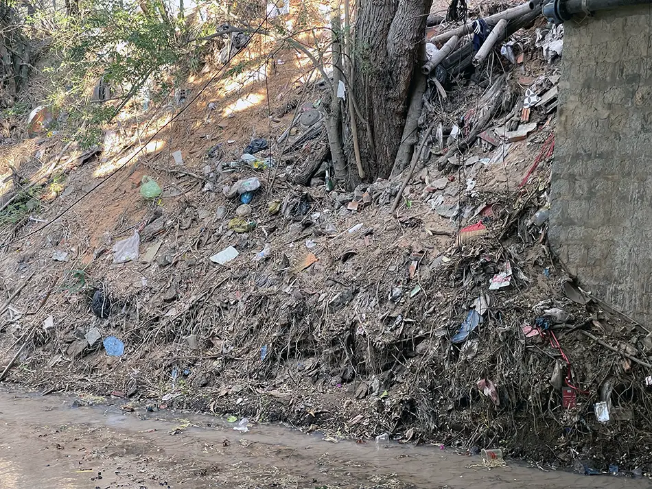 Plastic waste and debris accumulated along the bank at the lower entrance section of Fairy Stream, next to deteriorating sand-filled construction bags