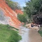 Two visitors wading through the shallow grey stream at Fairy Stream Mui Ne, with layered red and white sandstone cliffs rising on the left and a small vendor stall under palm trees on the right