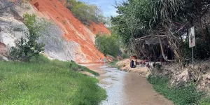 Two visitors wading through the shallow grey stream at Fairy Stream Mui Ne, with layered red and white sandstone cliffs rising on the left and a small vendor stall under palm trees on the right