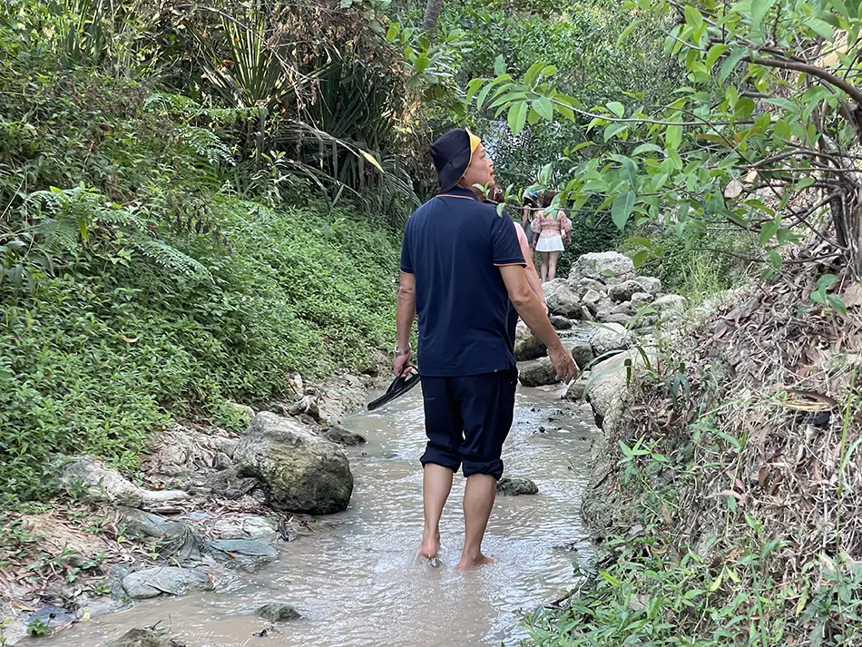 A visitor wading barefoot through ankle-deep grey water along the rocky mid-section of Fairy Stream, surrounded by dense tropical vegetation