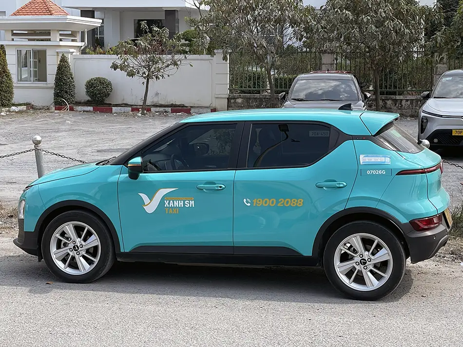 A turquoise electric taxi parked on a sandy roadside in Mui Ne with white resort buildings in the background