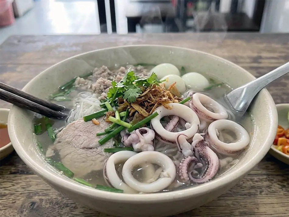 Bowl of hủ tiếu mực squid noodle soup with thin translucent noodles, squid rings, quail eggs, and fresh herbs