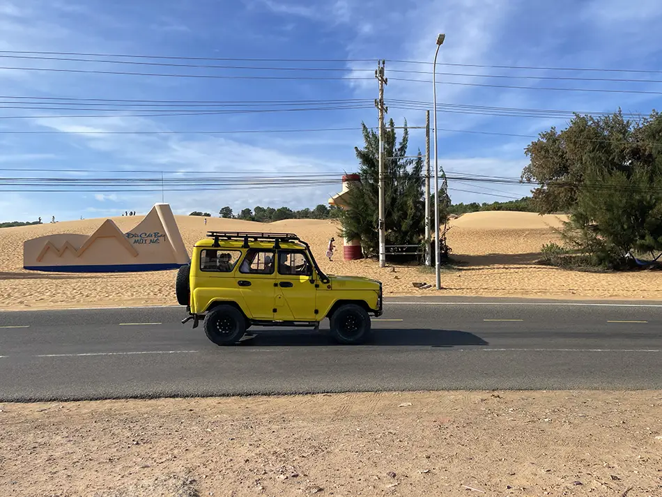 A yellow open-top jeep driving along a coastal road past the Red Sand Dunes in Mui Ne — the standard vehicle for backpacking Mui Ne sunrise tours