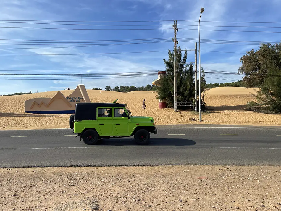 Green tour jeep driving past the Mui Ne red sand dunes on the main coastal road, with dune slopes rising directly behind