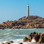 Ke Ga lighthouse rising above granite rock formations in rough sea, viewed from the mainland cape with waves breaking against the rocks