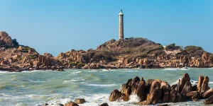 Ke Ga lighthouse rising above granite rock formations in rough sea, viewed from the mainland cape with waves breaking against the rocks