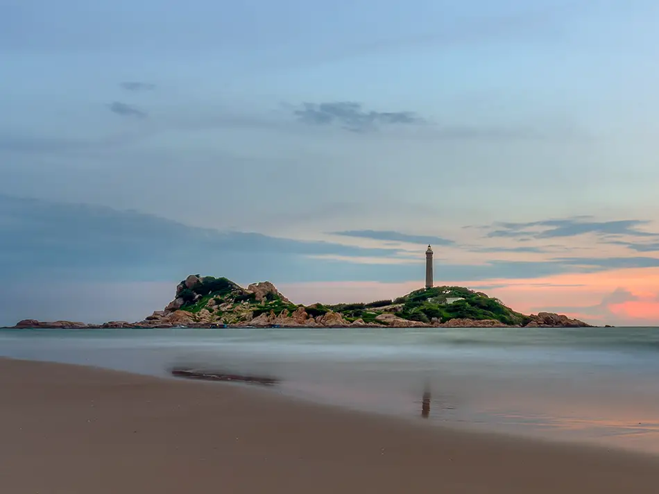 Ke Ga lighthouse on its rocky island silhouetted against a pastel pink and orange sunset sky, viewed across a calm stretch of beach