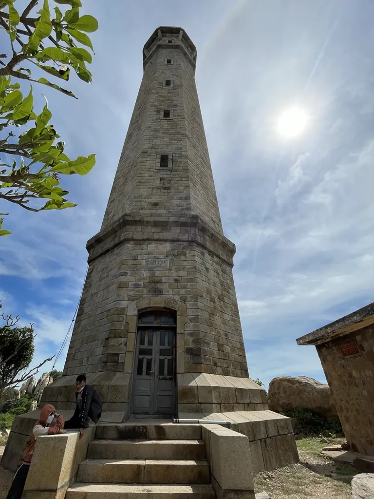 Close-up view of the tall narrow granite lighthouse tower at Ke Ga Cape with visitors at its base and tropical vegetation alongside
